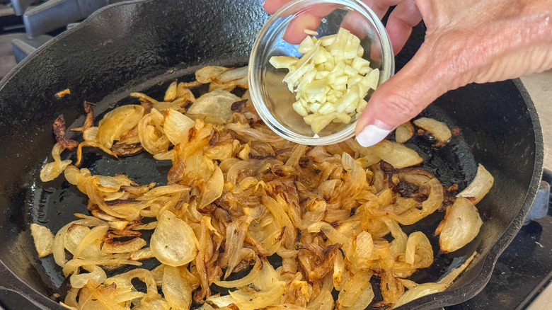 Hand adding small bowl of garlic to pan of onions