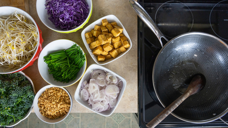 Overhead view of a HexClad pan on a stovetop, with a wooden spoon resting in the pan, and prepared ingredients for pad thai