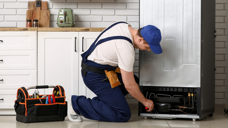 A repairman in a uniform inspecting the back of a refrigerator and repairing it with tools