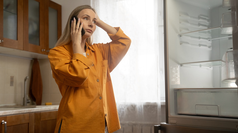 A woman standing in front of an empty, open refrigerator while on the phone with tech support
