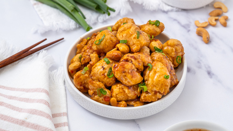 a dish of cashew chicken in a white bowl garnished with scallions with chopsticks and ingredients on the side.