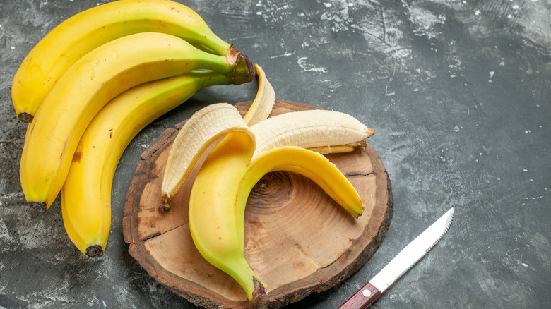 bunch of bananas next to a half-peeled banana on a wooden circle with a knife