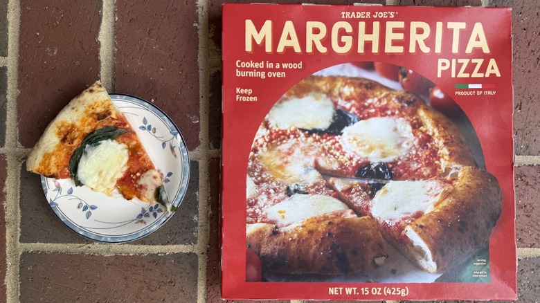 Slice of Trader Joe's Margherita pizza topped with mozzarella and several basil leaves, alongside box on brick surface