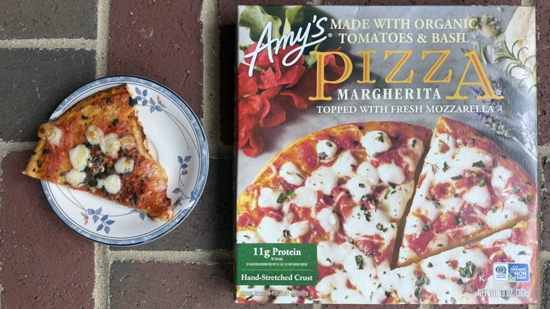 Slice of Amy's Margherita pizza topped with mozzarella and basil flecks on plate, alongside box on brick surface