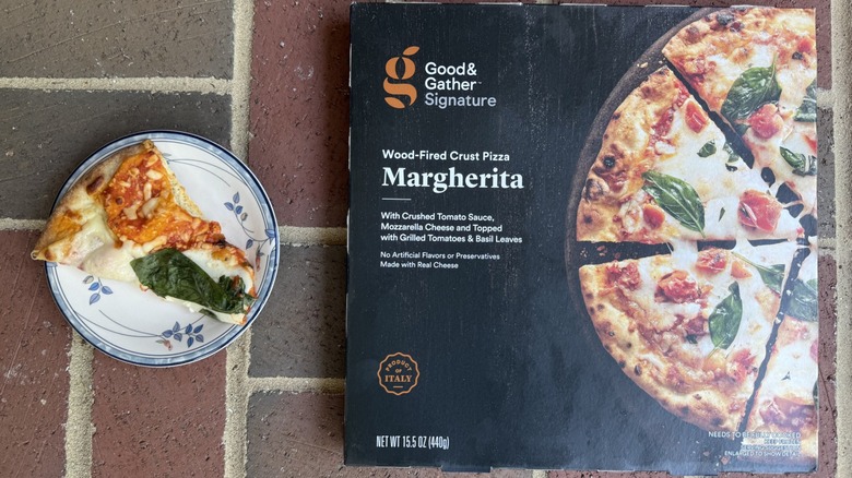 Slice of Good & Gather Margherita pizza topped with large basil leaf and mozzarella on plate, alongside box on brick surface
