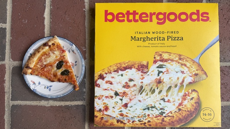 Slice of bettergoods Margherita pizza topped with two small basil leaves on plate, alongside box on brick surface