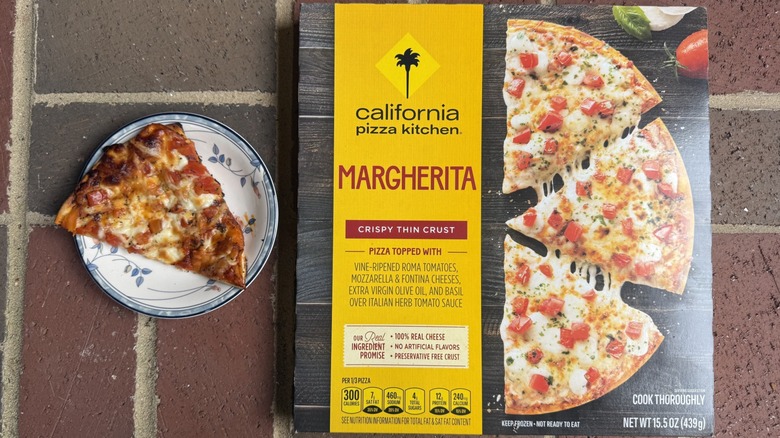 Slice of California Pizza Kitchen Margherita pizza topped with tomatoes and mozzarella on plate, alongside box on brick surface