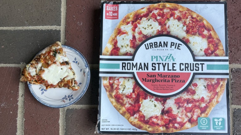 Slice of Urban Pie Margherita pizza with large blob of cheese on plate, alongside box on brick surface