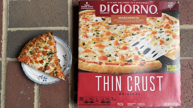Slice of DiGiorno Margherita pizza topped with several chunks of tomatoes and flecks of basil on plate, alongside box on brick surface