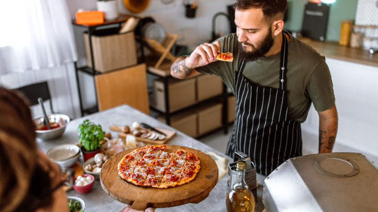 A man in an apron in his kitchen trying pizza someone is holding on a pizza peel
