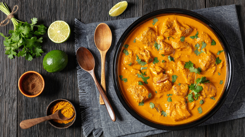 Curry on table alongside spoons, limes, cilantro, and spice powders