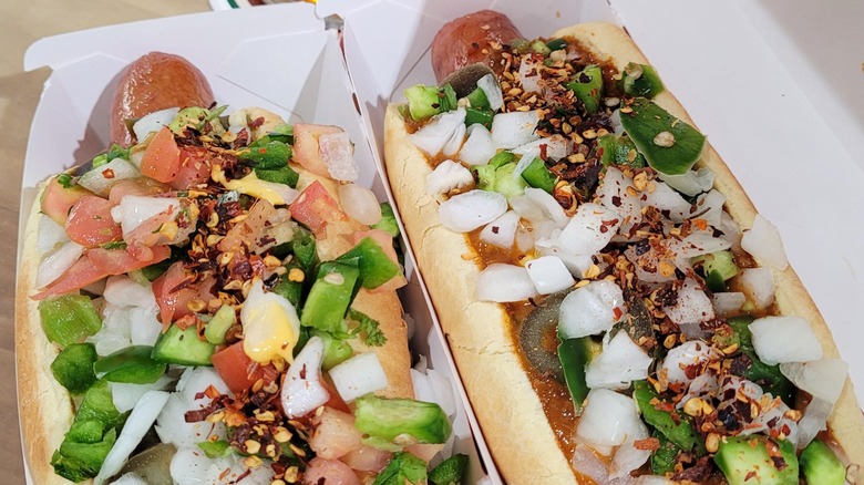 Two dhog dogs in cardboard trays with onions, peppers, and tomatoes