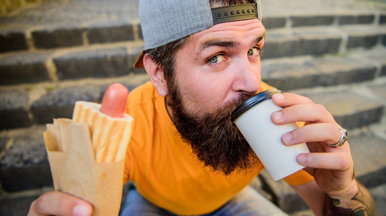 Guy sitting on steps outside drinking coffee and holding a hot dog