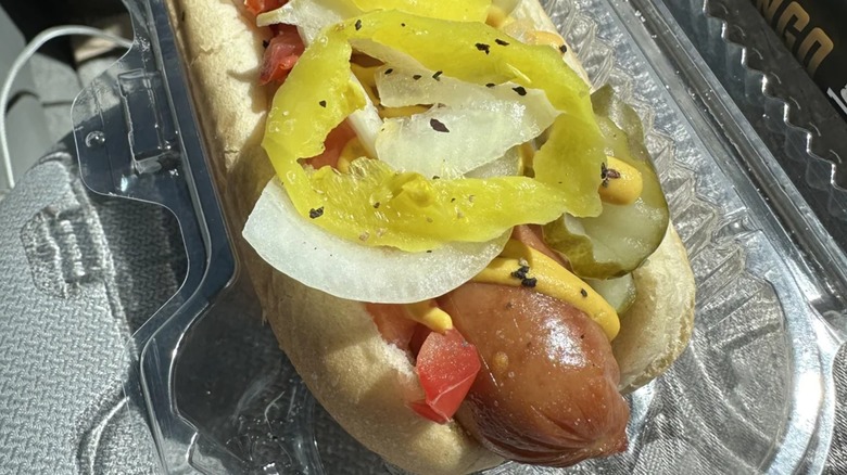 Hot dog in plastic container with onions, pickles, and banana peppers
