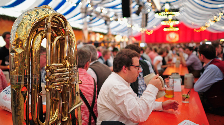 People drinking beer at tables under a German beer garden tent, with a tuba in the foreground