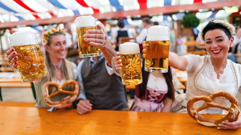 Four people raising glasses of beer, with two also holding pretzels, in a German beer garden tent
