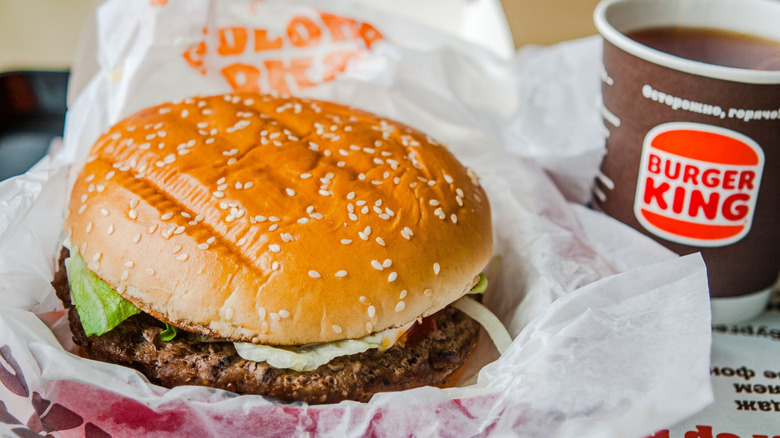 A Burger King burger sitting on the paper wrapping next to a cup of coffee