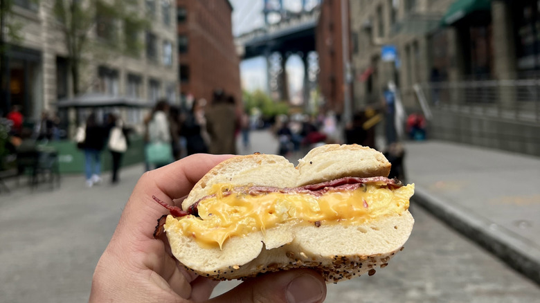 A hand holding a bacon, egg, and cheese sandwich, with a New York street and the Brooklyn Bridge in the background