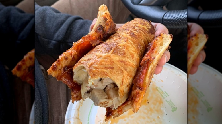 A person holding a slice of Costco pepperoni pizza wrapped around a food court Chicken Bake