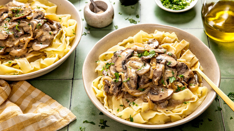 Two shallow beige bowls of beef stroganoff on table with other items.