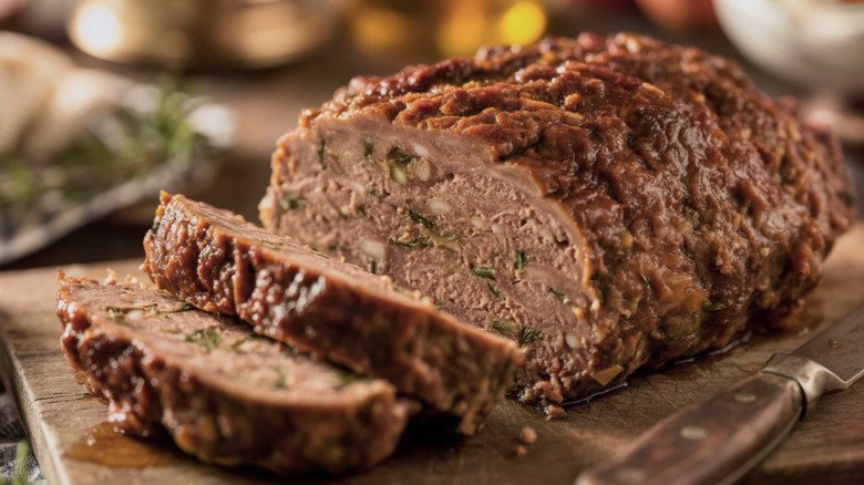 A meatloaf sits on a wooden cutting board next to a knife, with several slices of meatloaf already cut