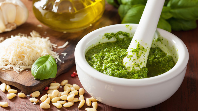 Freshly made pesto in a mortar with a pestle sits on a wooden table with other ingredients in the bcakground