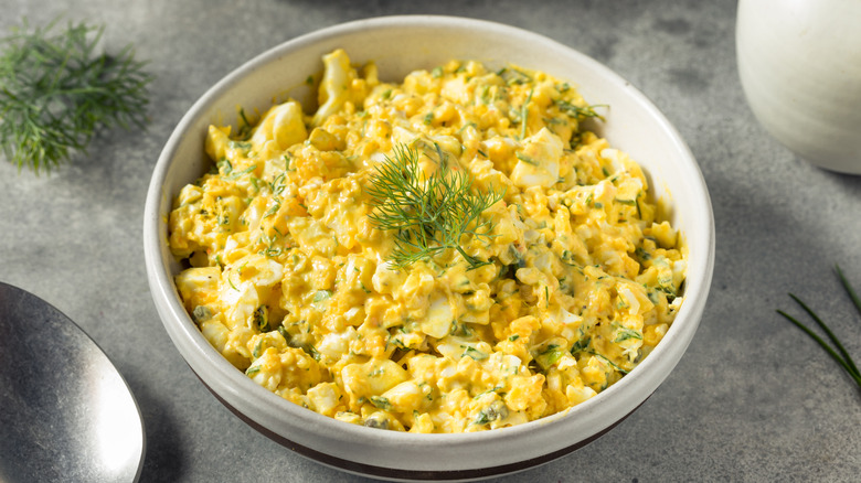 A bowl of egg salad on a grey work surface, with some fresh herbs and some other cutlery in the background