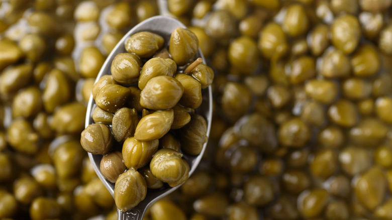 A metal spoon bearing a spoonful of pickled capers, held over many more capers beneath
