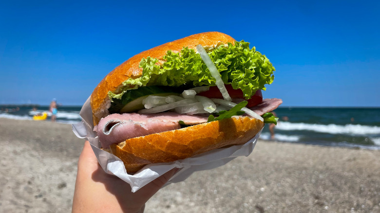 A hand holds a fischbrötchen filled with fish, lettuce, and onions on the beach.