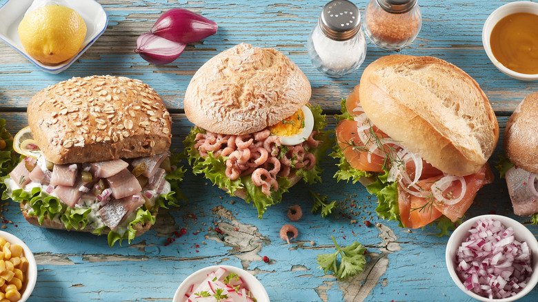 Three different types of fischbrotchen sit on a table with bowls of onion, radish, lemon, sauce, and salt and pepper shakers.