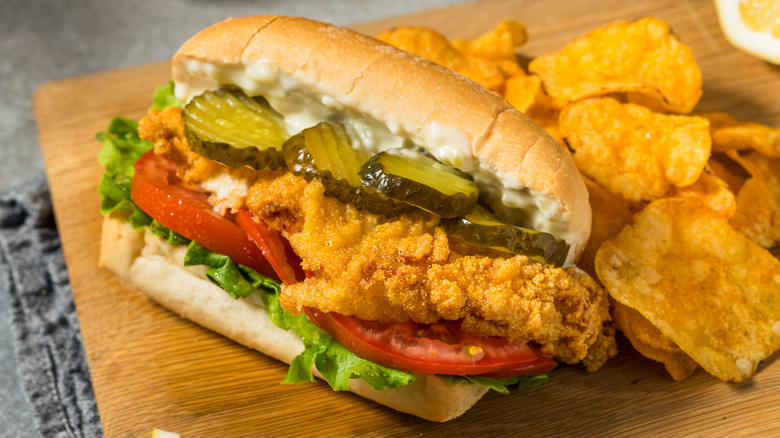 Closeup of a fried fish sandwich on a wood board with potato chips nearby.