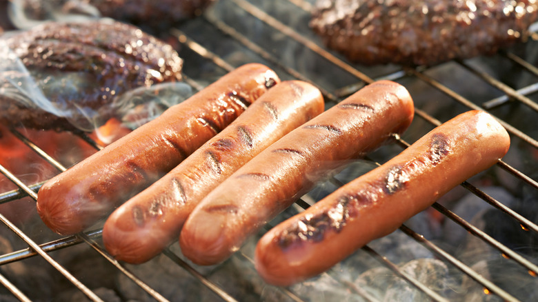 Four hot dogs cooking on a hot grill with burgers in the background
