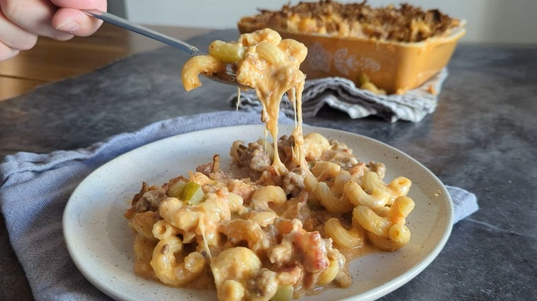 Someone holding a fork of Cowboy macaroni with bacon, spices, bell peppers and beef above a full plate, with casserole in the background.