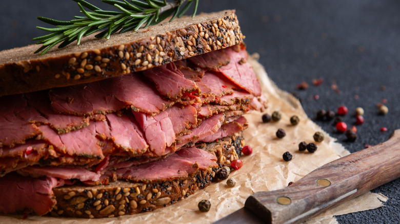 a pastrami sandwich with a sprig of rosemary on top sitting on wax paper beside a knife and loose peppercorns