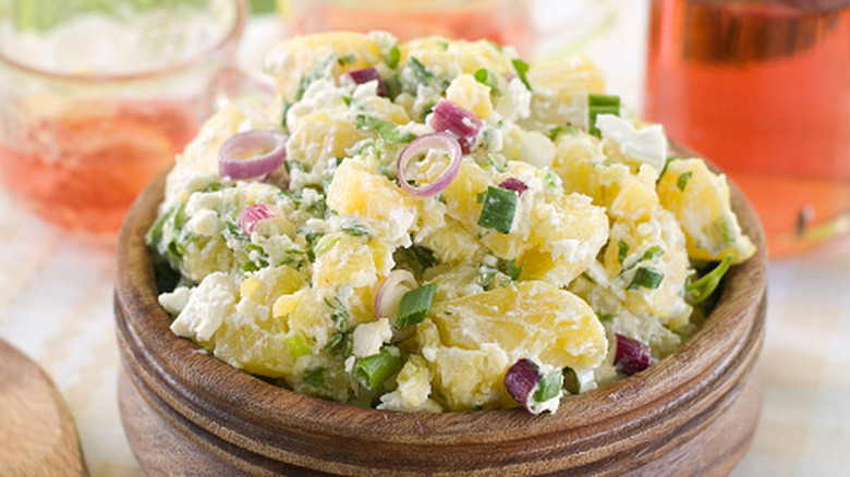 A wooden bowl of potato salad sitting on a table, with several glasses containing red liquid in the background