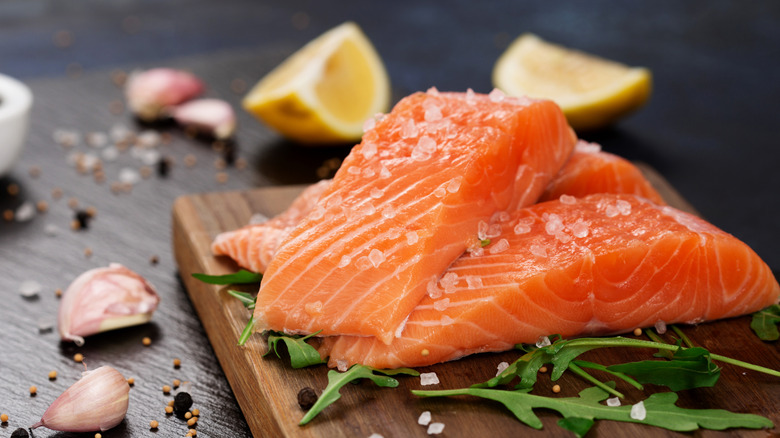 Fresh salmon fillets on a cutting board with arugula leaves and garlic cloves and lemon wedges in the background.