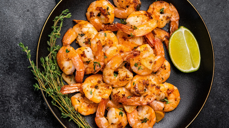 Black plate of shrimp scampi with lime, red pepper flakes, and thyme on black surface next to fork and cut limes