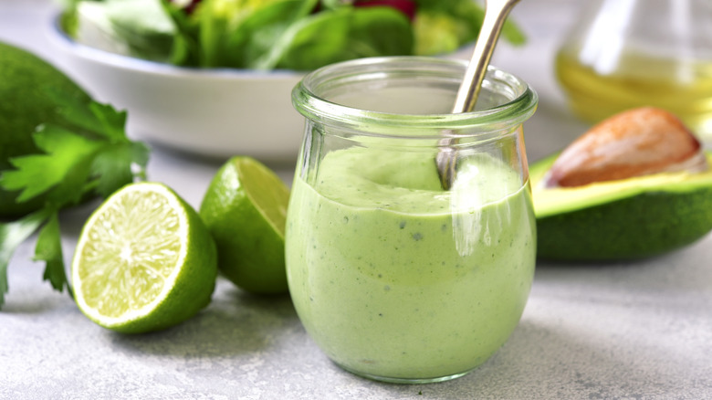 Avocado dressing in a glass serving next to lime halves and bowl of salad