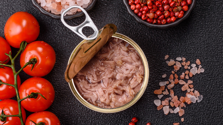 An open tuna can on a table beside cherry tomatoes, pink peppercorns, and pink salt