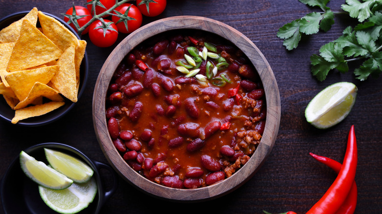 a wooden bowl of chile con carne surrounded by tomatoes, limes, tortilla chips, cilantro, and chiles
