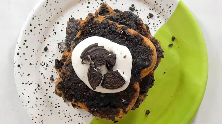 A stack of Oreo crumb-coated French toast, garnished with a cream cheese glaze and Oreo pieces, sitting on a white and green plate