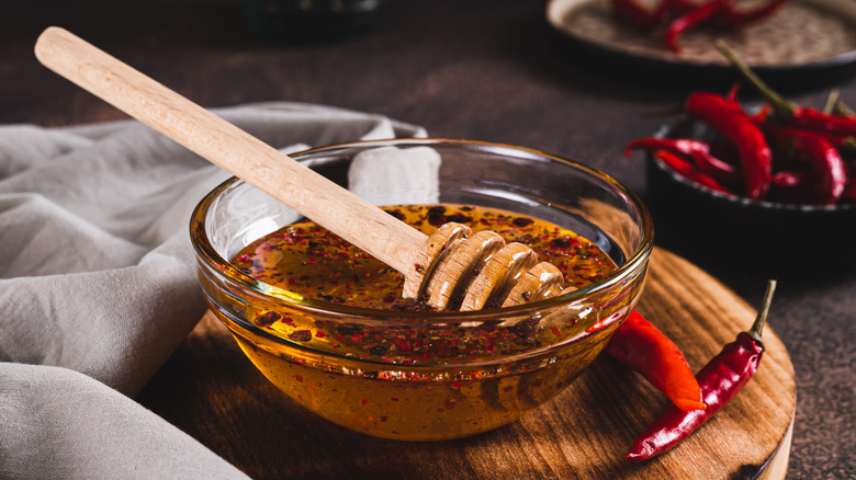 a glass bowl of hot honey with chili flakes on a wooden cutting board with chiles around it