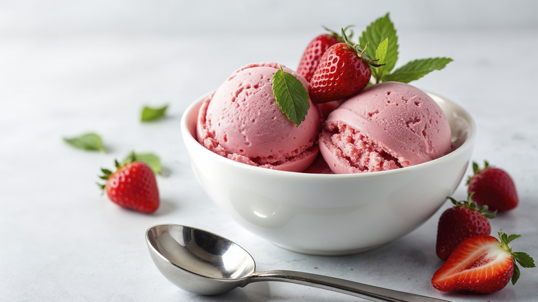 A white bowl with two scoops of strawberry sherbet topped with fresh strawberries and next to a metal spoon and a few scattered strawberries