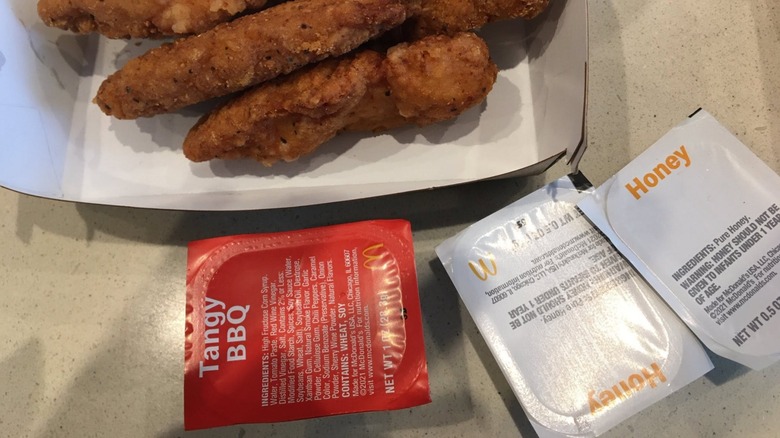Chicken strips in a cardboard container above a package of McDonald's Tangy BBQ sauce and two packages of McDonald's honey