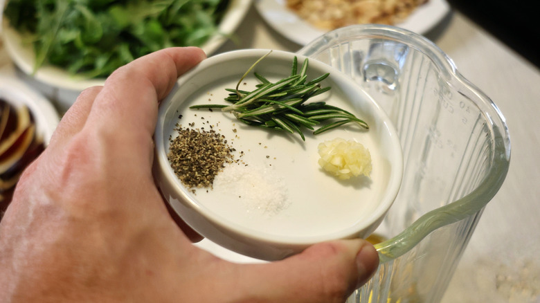 hand holding dish of salt, pepper, minced garlic, and rosemary leaves above a blender