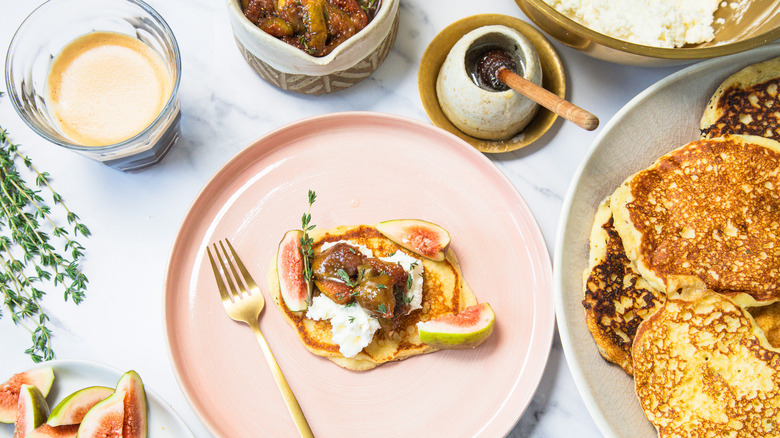 A table scene of one goat cheese pancake on a plate with all the toppings and pancakes around.