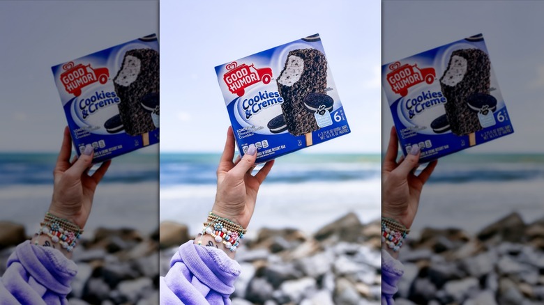 Hand holding box of Good Humor Cookies and Cream Bar outside on a rocky beach.