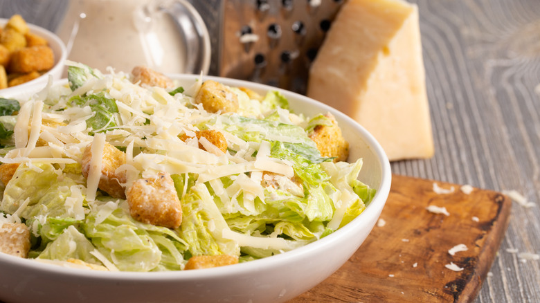 A caesar salad with fresh parmesan cheese and croutons is in a bowl on a cutting board.