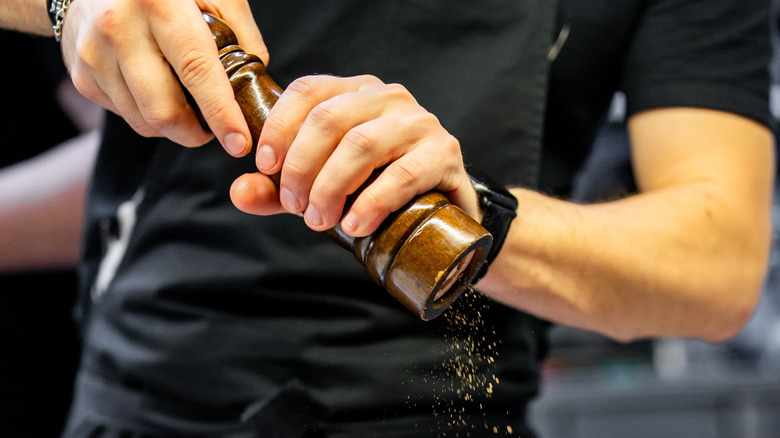 cook in T-shirt and black apron twists a classic-style wooden pepper mill, with fresh-ground pepper cascading from the grinder mechanism