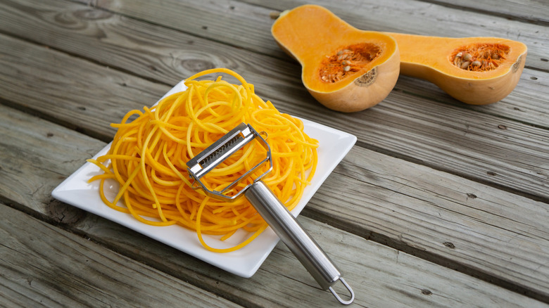 julienne peeler shown leaning on a square white plate filled with julienned squash "noodles," with a halved butternut squash behind it